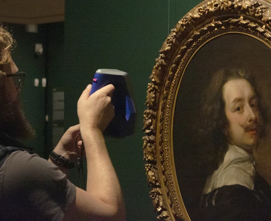 A man using a handheld 3D scanning device on a framed painting in a museum, capturing the artwork’s details.