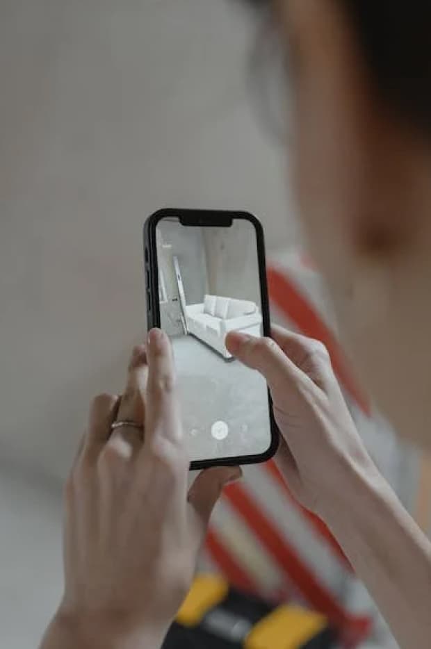 A person's hand holding a smartphone, viewing an augmented reality (AR) model of a white sofa in a room, demonstrating the use of AR in interior design.