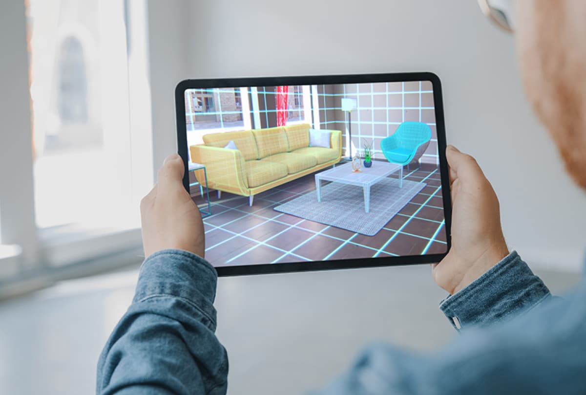 A person's hand holding a tablet displaying an augmented reality view of a modern living room with a yellow sofa, white coffee table, and a blue chair, superimposed on a grid overlay.