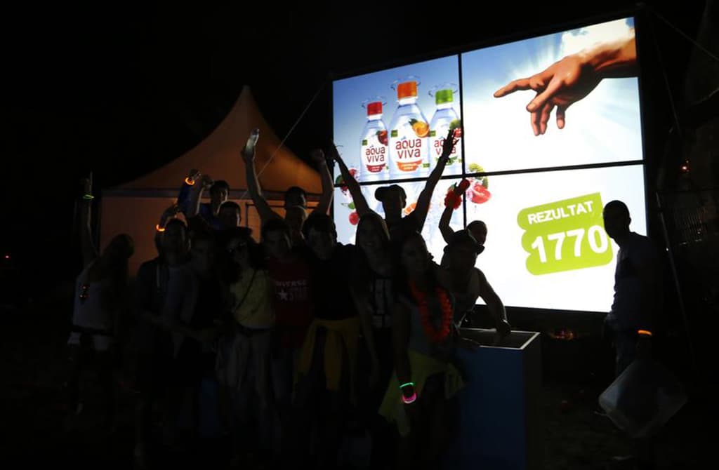 A group of people raising their hands in excitement at night, in front of a large screen displaying a promotional image with a hand pointing to a product and text that seems to indicate a score or result.