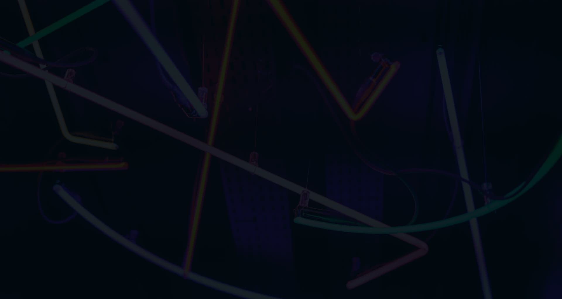 An abstract, dark background with neon lines in various colors creating a network of geometric shapes and angles, giving the impression of a modern, artistic light installation or the visualization of a complex, interconnected system.