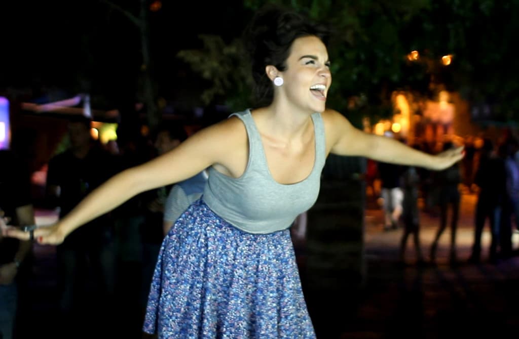 A joyful woman with her arms outstretched, spinning around at an outdoor nighttime event. The background is softly lit and blurred, highlighting her smiling expression and the motion of her twirling skirt.