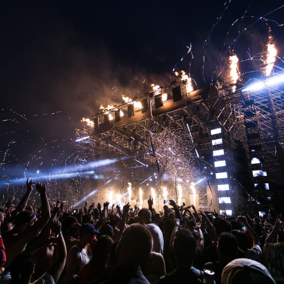 Music festival. Concert crowd with raised hands enjoying a live music performance under a night sky, with stage lights and pyrotechnics adding to the festive atmosphere.