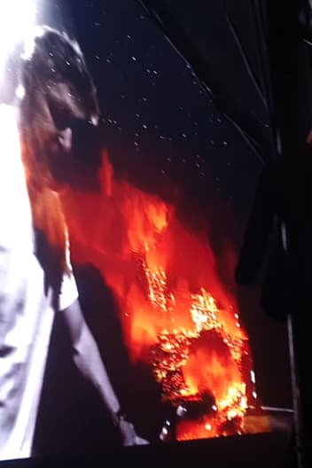 A person in the foreground with a dynamic background displaying what appears to be flames, possibly from a video or projection