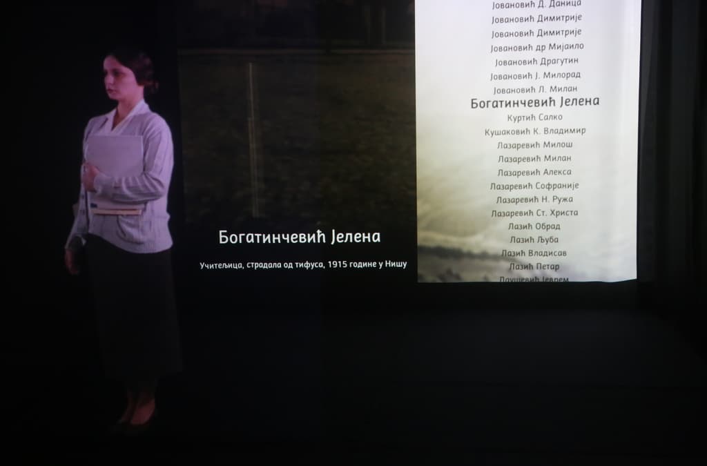 A life-size holographic projection of a historical figure in period clothing, standing beside a wall list of names, part of an educational exhibit or memorial presentation.