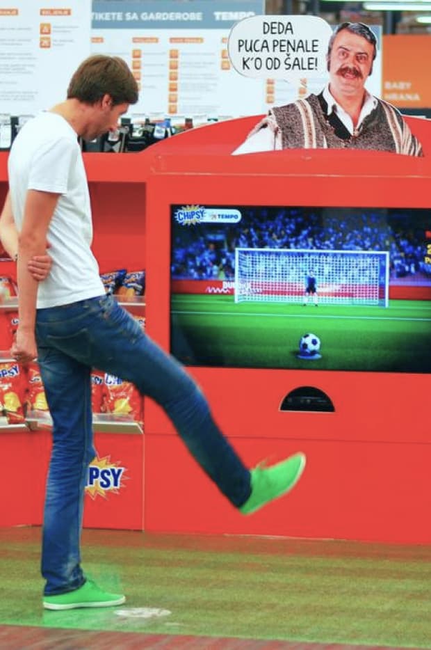 A person playfully kicking towards a virtual soccer goal displayed on a digital screen at a promotional event, simulating a real-life penalty kick.