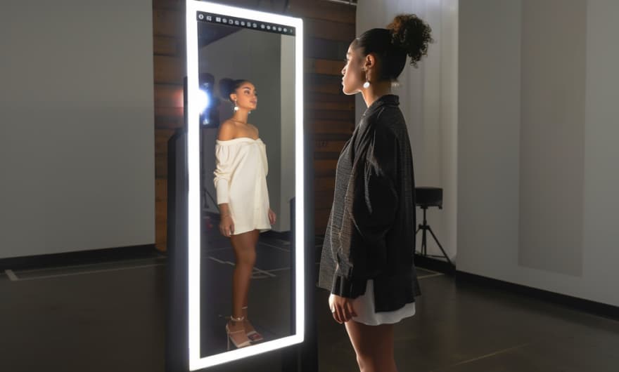 A woman standing in front of a full-length smart mirror that displays a virtual fitting of her in a white dress, reflecting both her actual appearance and the digital outfit.