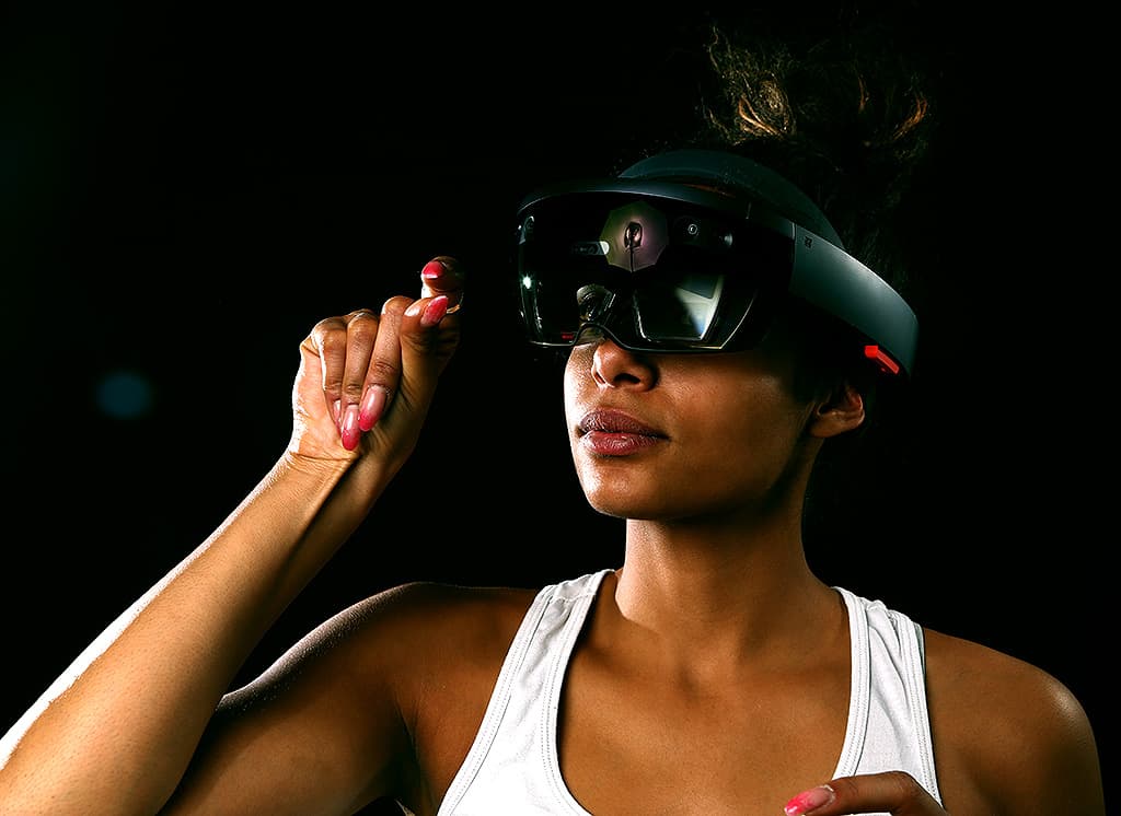 A woman with her hair pulled back, wearing an augmented reality headset, lifts her hand to adjust the device, set against a dark background.