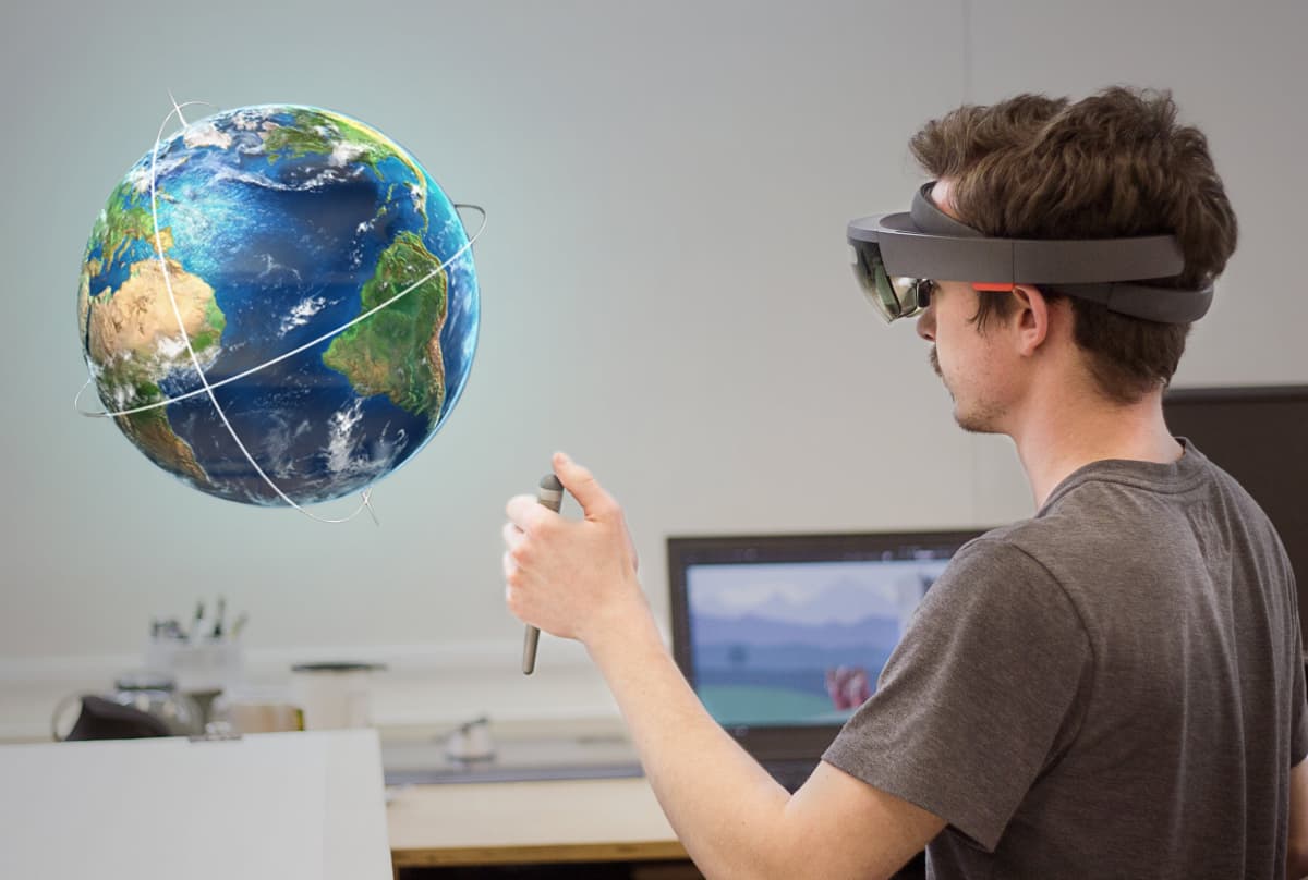 A person wearing augmented reality glasses interacting with a 3D holographic model of Earth in a room with a monitor displaying a mountain landscape in the background.