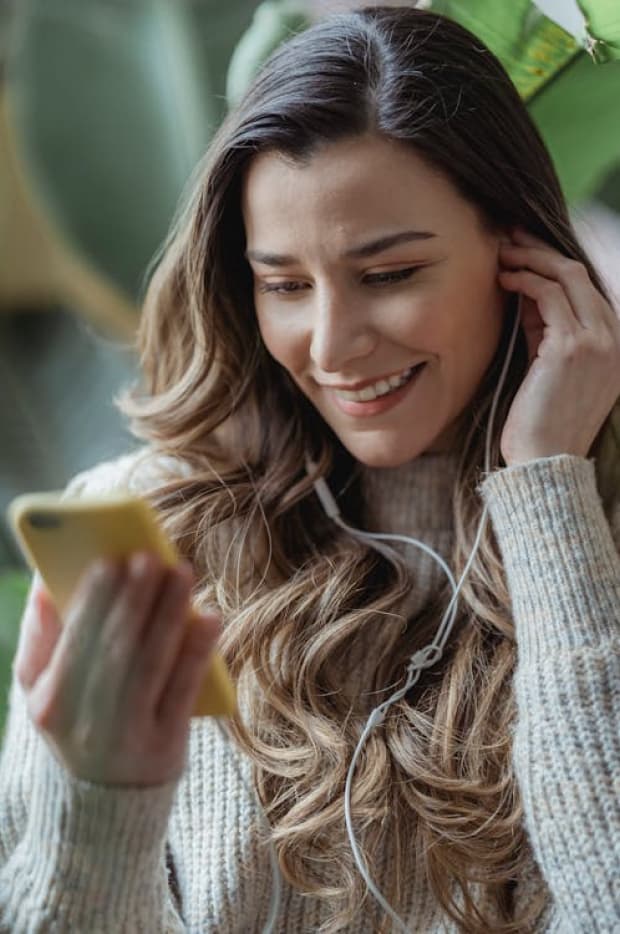 A smiling woman with headphones looking at her smartphone, her expression reflecting enjoyment and engagement with the content she is viewing.