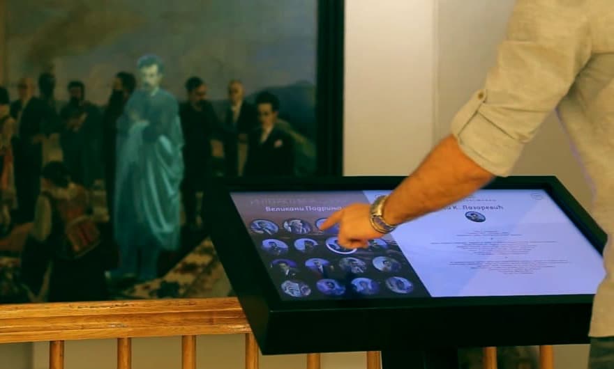 A man interacting with an interactive touchscreen display in an exhibition space, with a large, classical painting in the background.