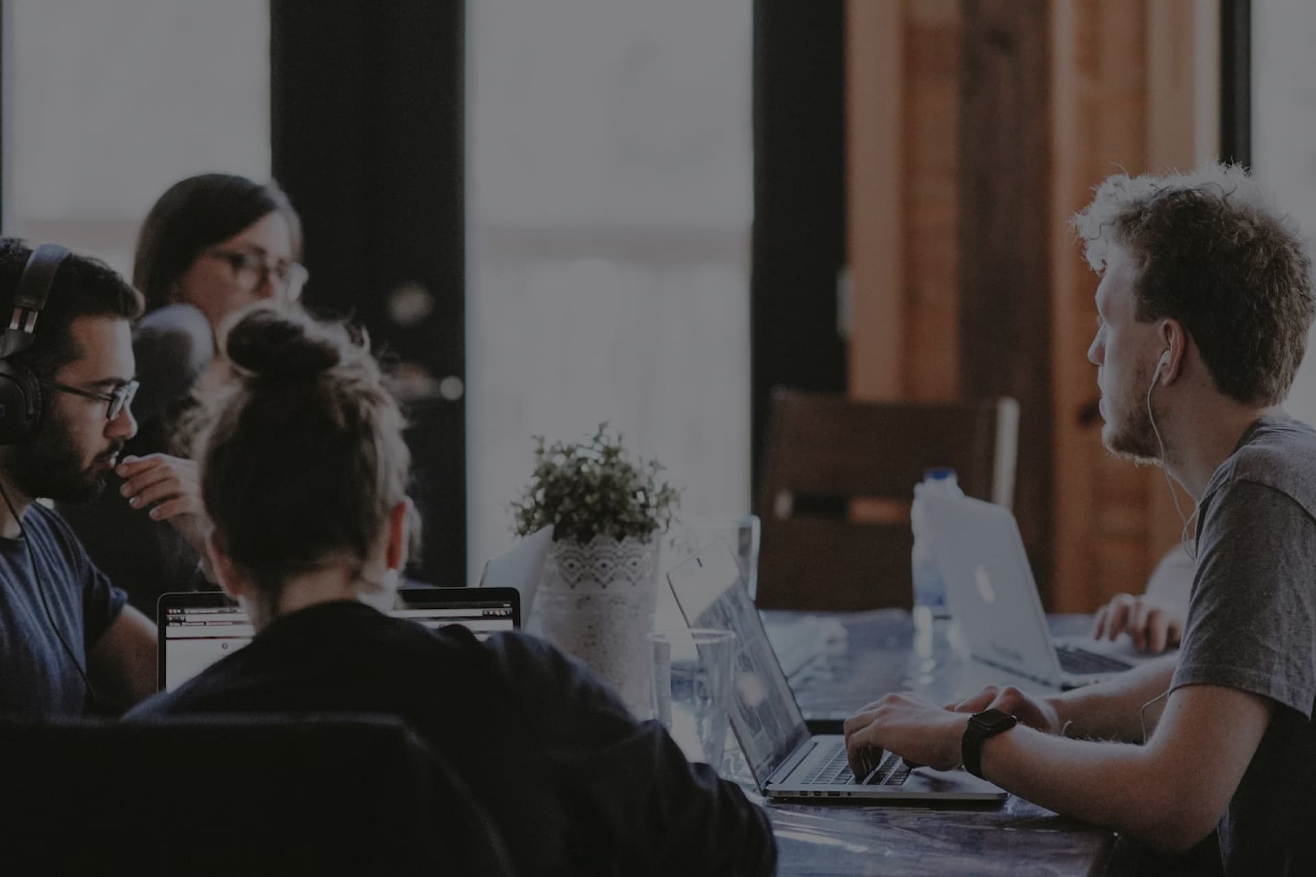 A team of professionals in a collaborative work session, engaged in discussion and work on laptops, in a casual office setting.