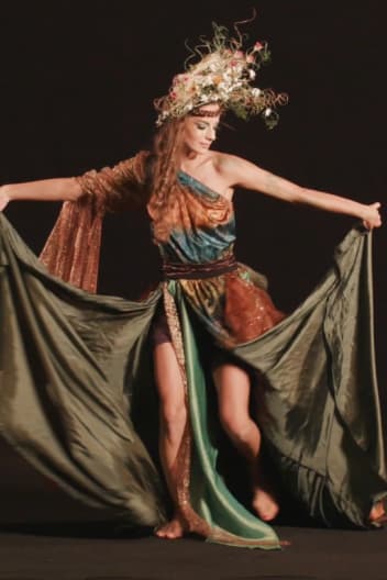 A dancer in ornate costume with a headdress performs, arms extended, against a black background.
