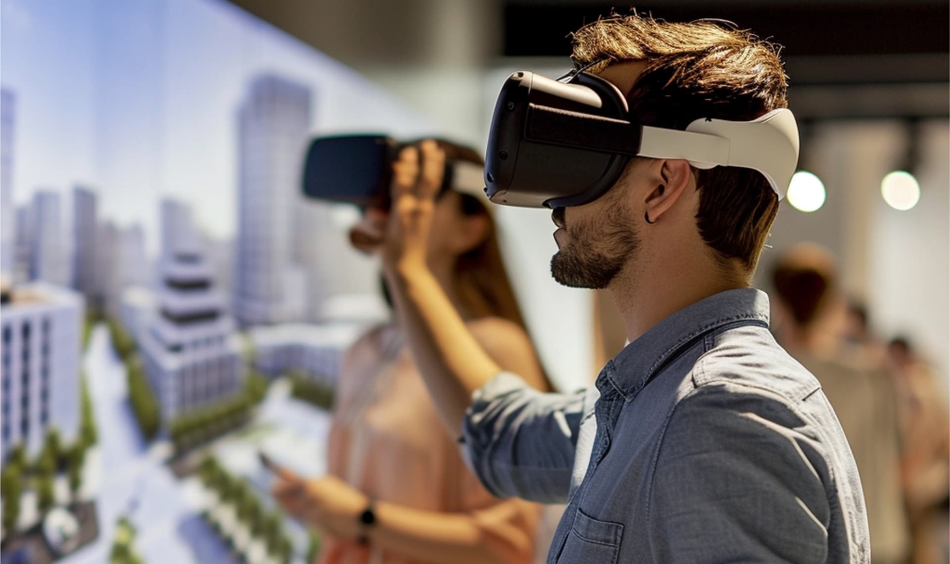 A man and a woman in casual clothing standing indoors using virtual reality headsets, with blurred images of a cityscape model in the background, suggesting a virtual interaction with a three-dimensional environment.