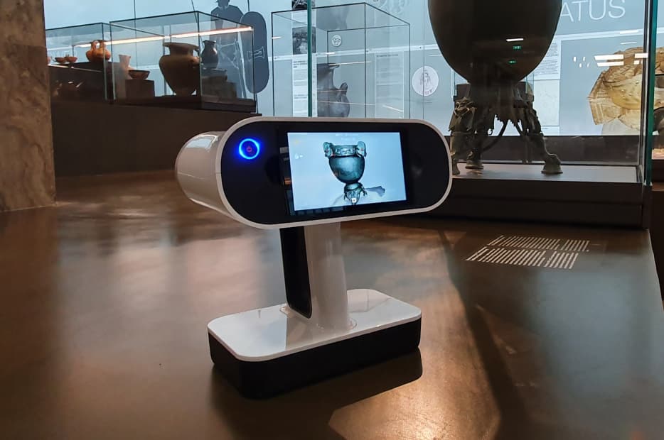 A modern robotic information kiosk with a digital display screen, showing a blue-eyed humanoid robot, situated in a museum with transparent glass walls and artifacts in the background.