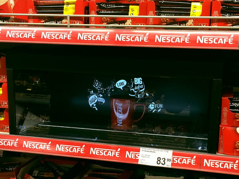 An interactive digital shelf display in a retail store, with a virtual model of a NESCAFÉ coffee jar surrounded by physical product packaging, demonstrating a blend of physical and augmented reality marketing.