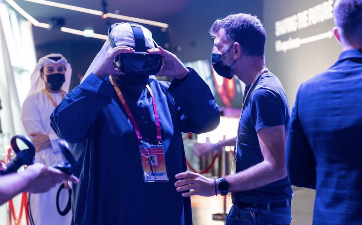 A person wearing a VR headset is assisted by a technician at a high-tech event with diverse attendees and ambient lighting.