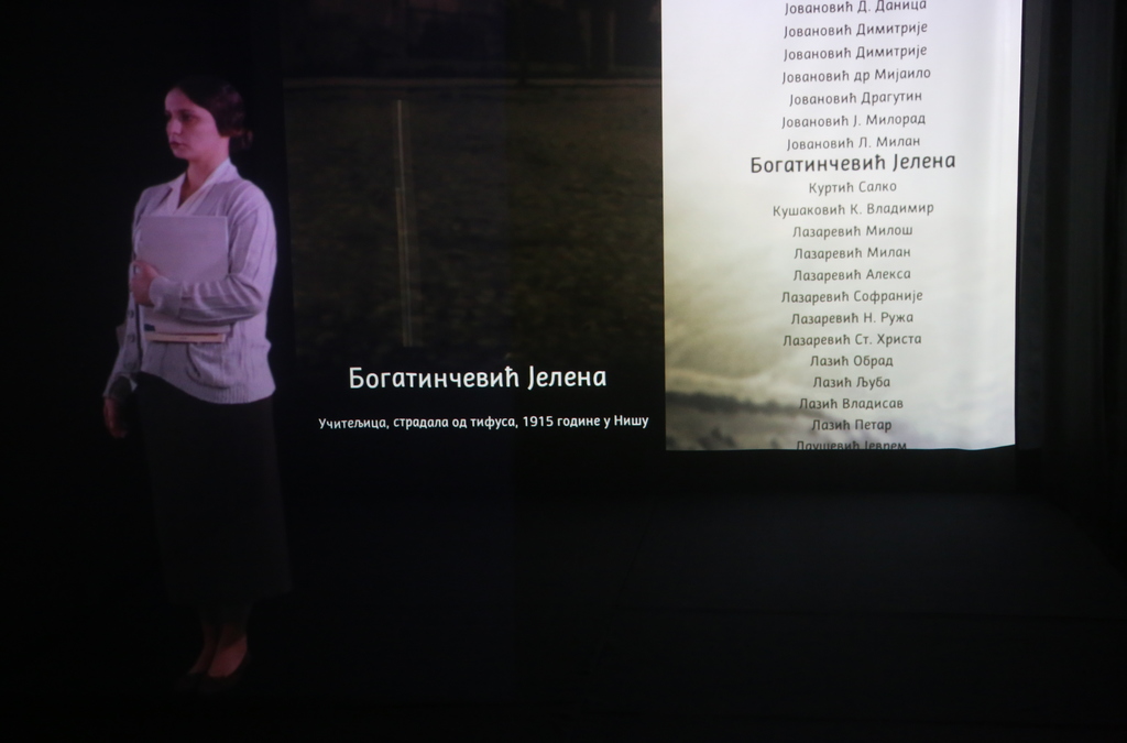 A life-size holographic projection of a historical figure in period clothing, standing beside a wall list of names, part of an educational exhibit or memorial presentation.